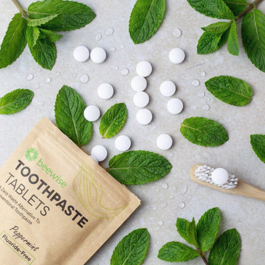 Draufsicht auf weiße beewise Zahnpasta-Tabletten, die aus einer Papiertüte auf eine helle Steinfläche neben frischen grünen Minzblättern verstreut liegen.