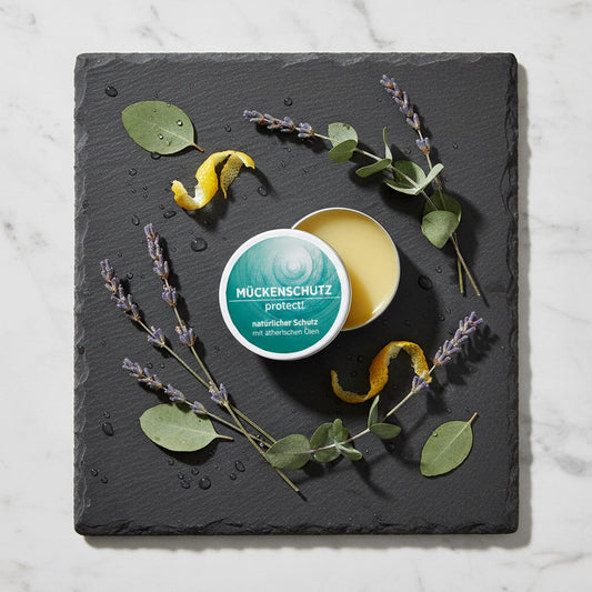 Die geöffnete Dose des Sonnengrün Mückenschutzes auf einer Schieferplatte, dekoriert mit Lavendel, Eukalyptusblättern und Zitronenschale zur Darstellung der natürlichen Inhaltsstoffe.