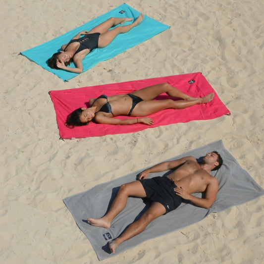 Drei Personen liegen am Strand auf farbenfrohen OBABA Strandmatten in Grau, Pink und Türkis – kompakte Reisebegleiter aus Bio-Baumwolle.