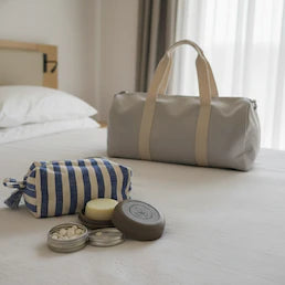 Eine graue Reisetasche, ein Kulturbeutel und nachhaltige Reiseartikel wie festes Shampoo liegen auf einem Hotelbett, passend zur trekaro-Kollektion "Reiseessentials".