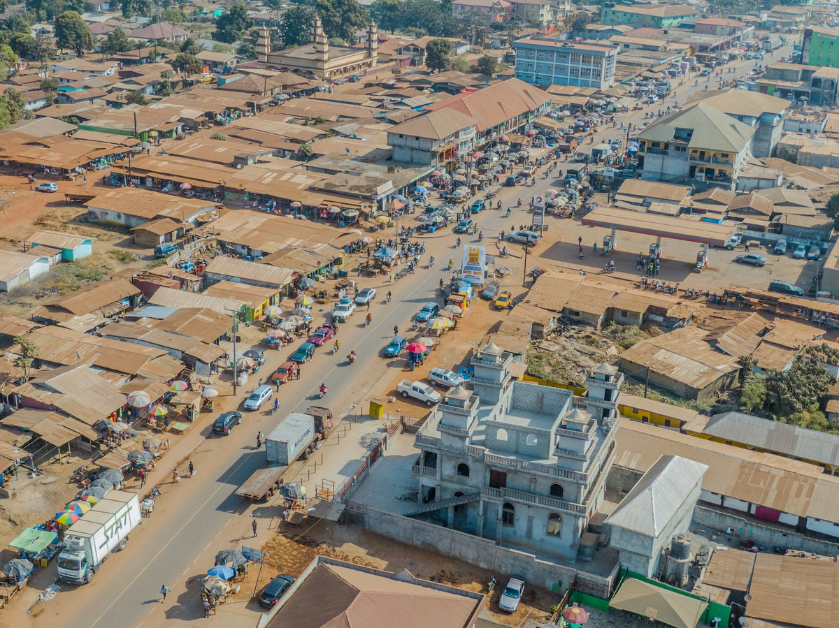 Vogelperspektive auf eine belebte Hauptstraße in Ganta, Liberia, mit dicht bebauten Häuserreihen, Marktständen, Fahrzeugen und einer Moschee im Vordergrund.