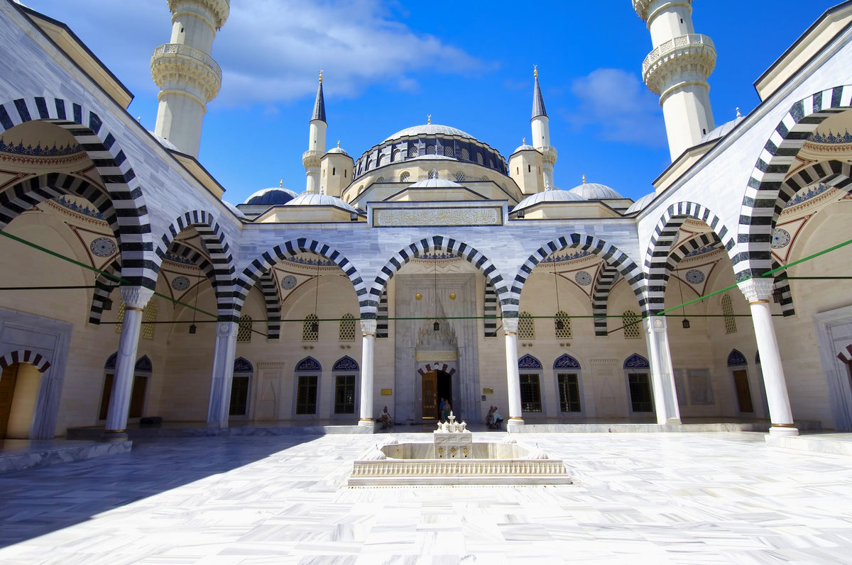 Sonnendurchfluteter, hell gepflasterter Innenhof der Hazrat-Omar-Moschee in Aschgabat mit schwarz-weiß gestreiften Bögen, Säulengängen, einem Brunnen im Zentrum und schlanken Minaretten.