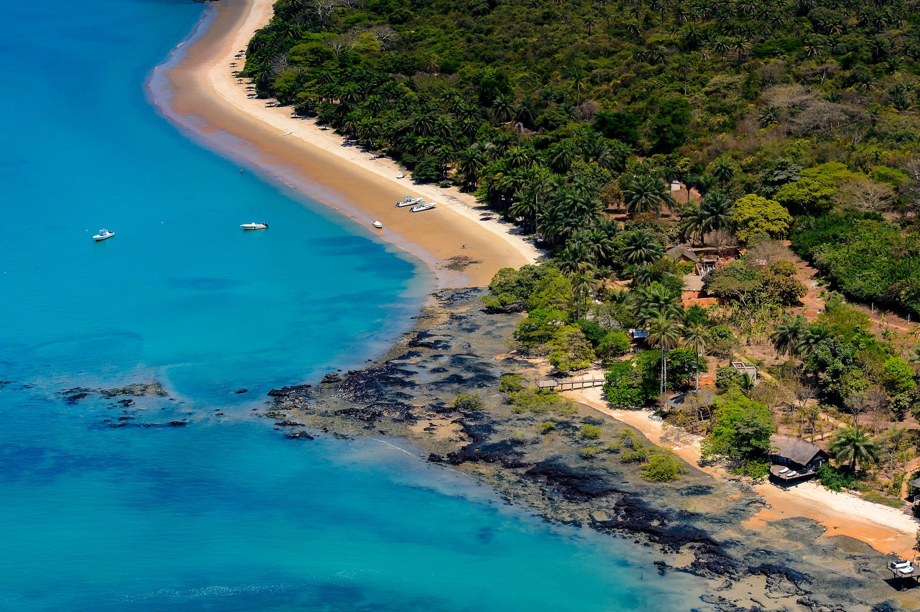 Luftaufnahme einer palmengesäumten Bucht mit goldgelbem Sandstrand, türkisfarbenem Meer und dunklen Felsformationen im flachen Wasser.