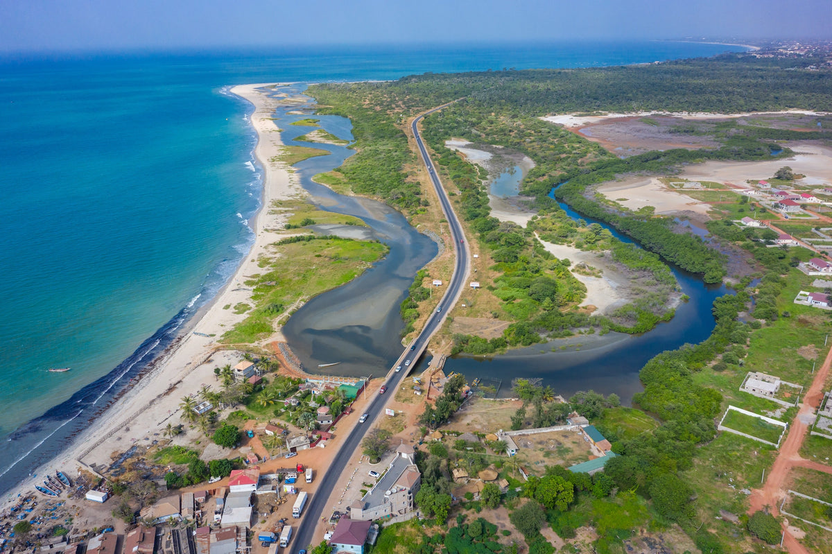 Luftaufnahme einer asphaltierten Küstenstraße in Gambia, die sich zwischen dem blauen Atlantik, einem Sandstrand und grünen Flussdeltas erstreckt.