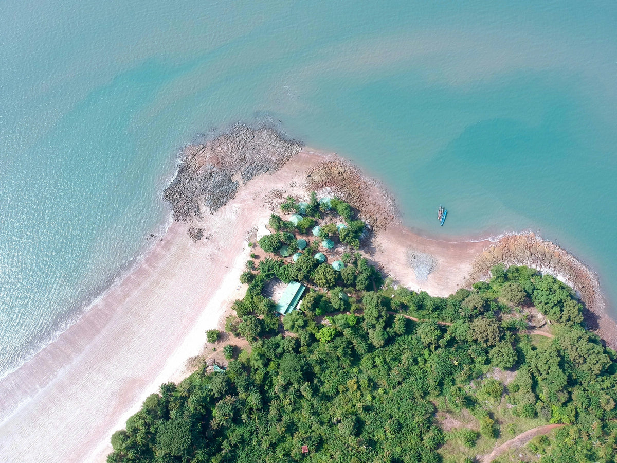 Senkrechte Drohnenaufnahme einer dicht bewaldeten Landzunge mit Sandstrand, kleiner Hüttensiedlung und zwei kleinen Booten im türkisfarbenen Meer.