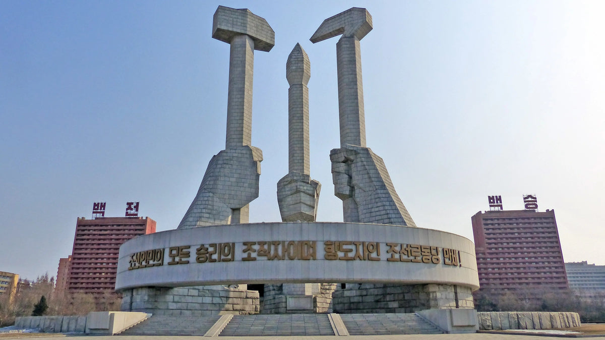 Monumentales Stein-Denkmal in Nordkorea, das drei riesige Hände zeigt, die einen Hammer, einen Pinsel und eine Sichel emporhalten, umgeben von einem kreisförmigen Bauwerk mit koreanischer Schrift.