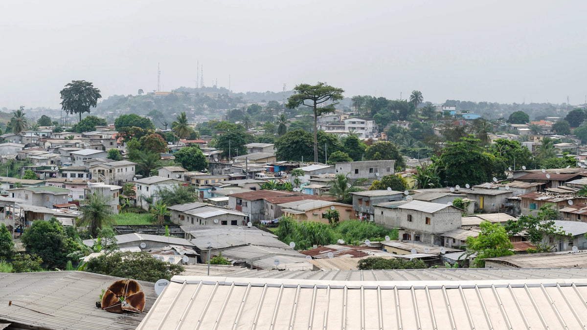 Ein breiter Panoramablick über ein dicht bebautes Wohnviertel in Libreville (der Hauptstadt von Gabun) mit eng stehenden Häusern, vielen Blechdächern und dazwischen wachsenden Bäumen unter einem bewölkten Himmel.