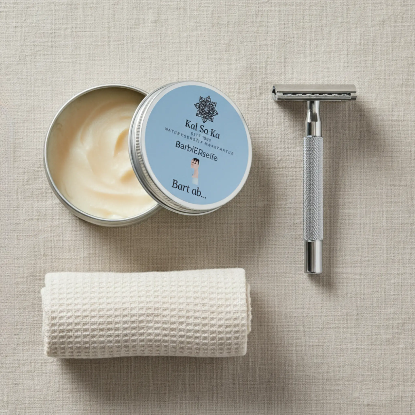 Draufsicht auf ein Rasur-Set: eine offene Dose Kal Sa Ka Barbierseife, ein silberner Rasierhobel und ein gerolltes, beiges Waffeltuch auf einem Leinenhintergrund.