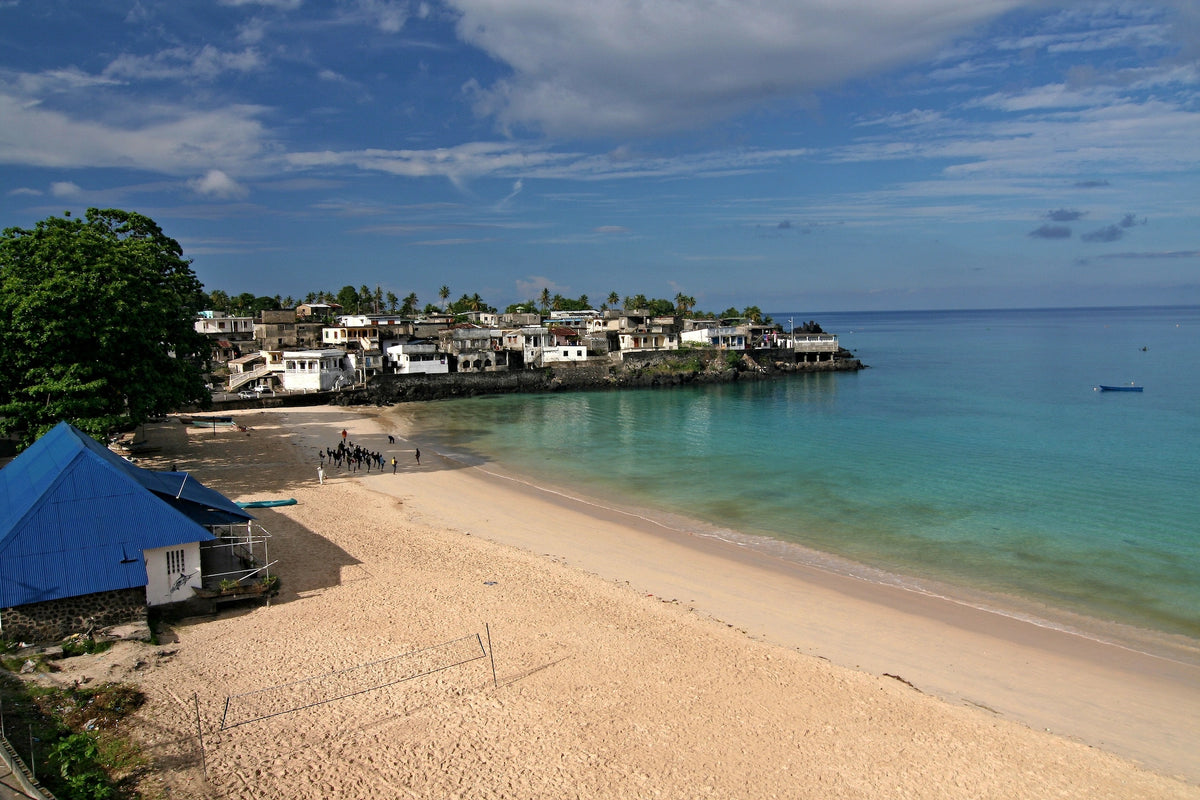 Sandy beach with a volleyball net, leading to turquoise ocean water and a small coastal village built on dark volcanic rock under a bright blue sky.