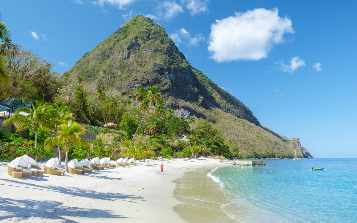 Malerischer weißer Sandstrand mit Palmen und Liegestühlen an einer ruhigen Bucht, überragt von dem steil aufragenden, grün bewachsenen Berg Petit Piton unter blauem Himmel.
