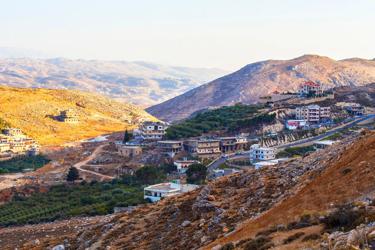 Weitwinkelaufnahme eines bergigen Tals im Libanon mit vereinzelten Häusern auf trockenen, sonnenbeschienenen Hügeln und einer Straße im Hintergrund.