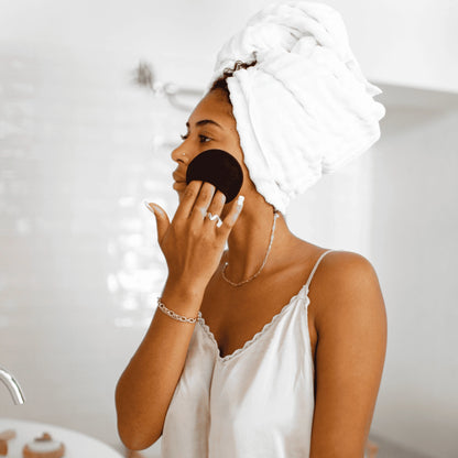 Eine Frau mit weißem Handtuch-Turban reinigt ihr Gesicht mit einem schwarzen, waschbaren Make-up-Entferner-Pad von Bamboovement vor dem Badezimmerspiegel.