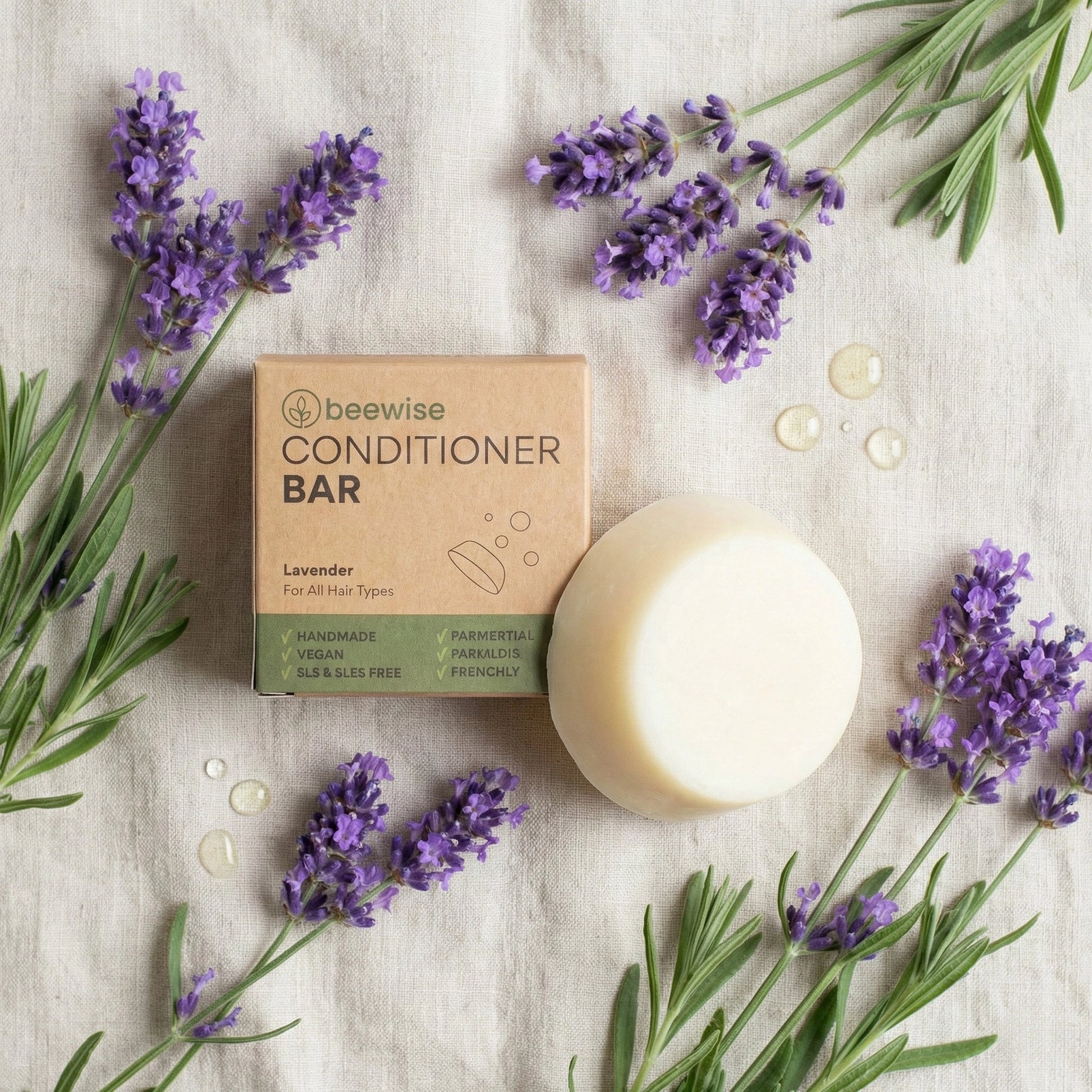Draufsicht auf den beewise Conditioner Bar Lavendel in seiner nachhaltigen Kartonverpackung, umgeben von frischen Lavendelzweigen und Öltropfen auf hellem Leinenstoff.