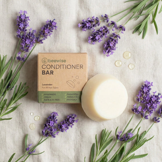 Draufsicht auf den beewise Conditioner Bar Lavendel in seiner nachhaltigen Kartonverpackung, umgeben von frischen Lavendelzweigen und Öltropfen auf hellem Leinenstoff.