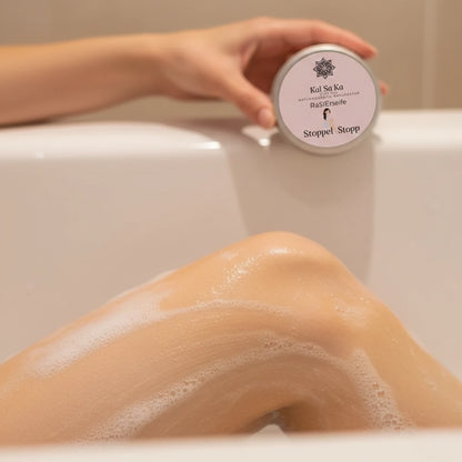 Anwendung der 'Stoppel Stopp' Rasierseife in der Badewanne: Eingeschäumtes Bein einer Frau im Vordergrund, im Hintergrund wird die Dose Naturkosmetik-Rasierseife gehalten.