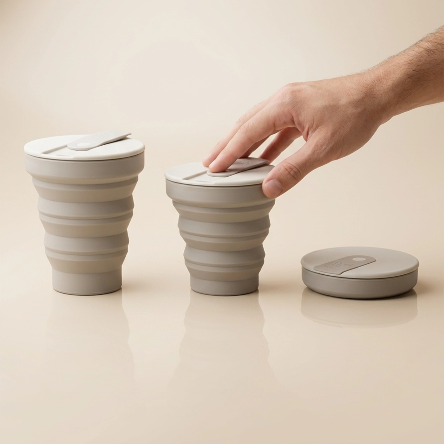 A hand presses down on a collapsible travel cup, next to another cup and a collapsed cup.