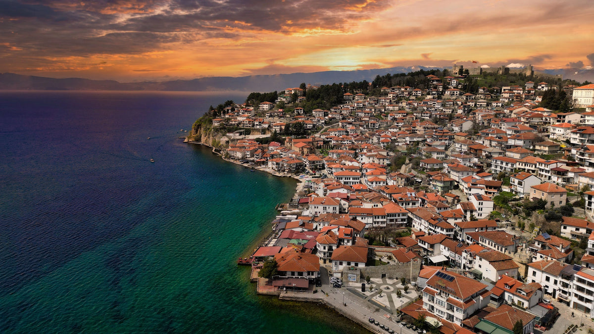 Luftaufnahme einer historischen Stadt mit rotdachigen Häusern an einem tiefblauen See bei dramatischem orangefarbenem Sonnenuntergangshimmel.