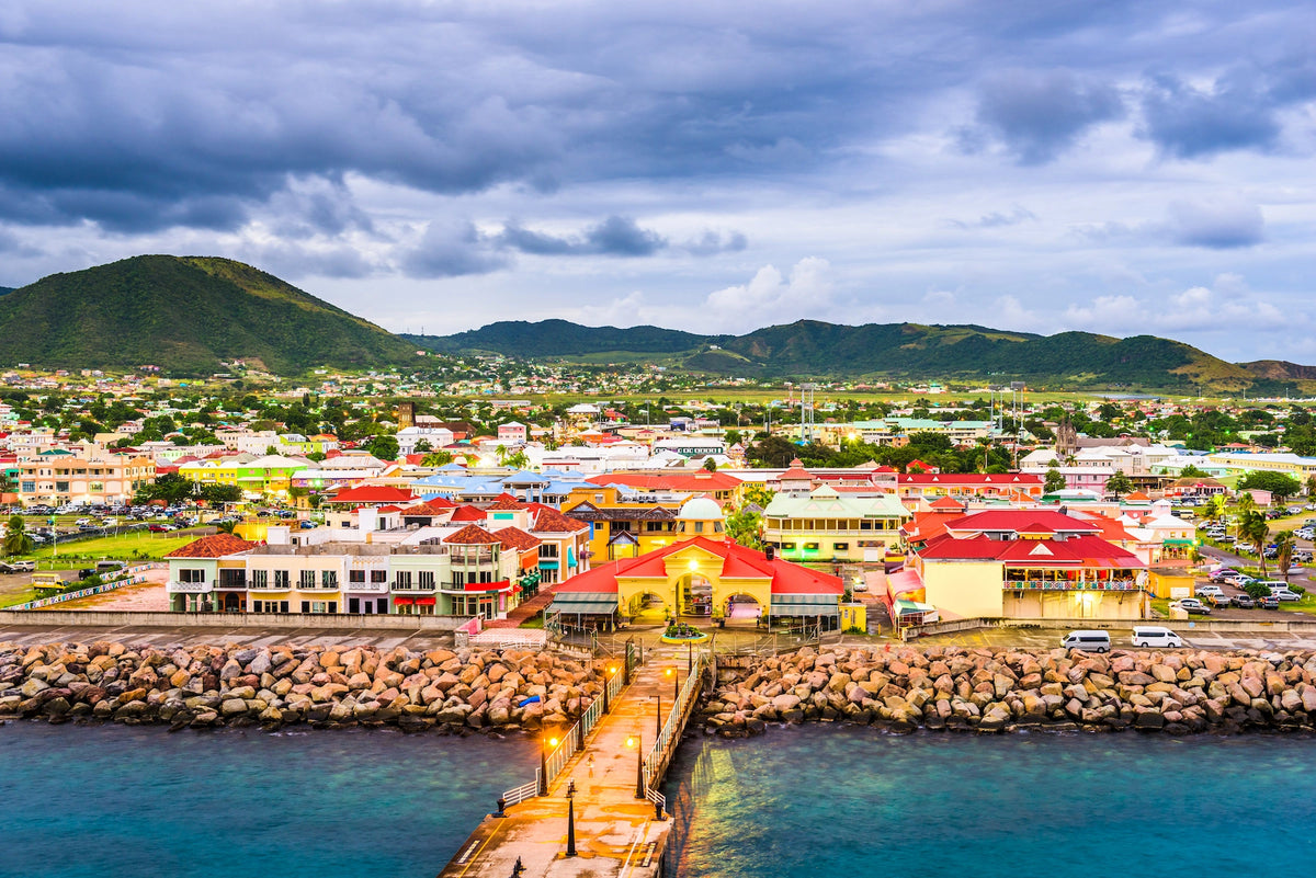 Blick vom Wasser auf die farbenfrohen Gebäude von Basseterre mit einem Steg im Vordergrund und grünen Hügeln unter leicht bewölktem Himmel im Hintergrund.