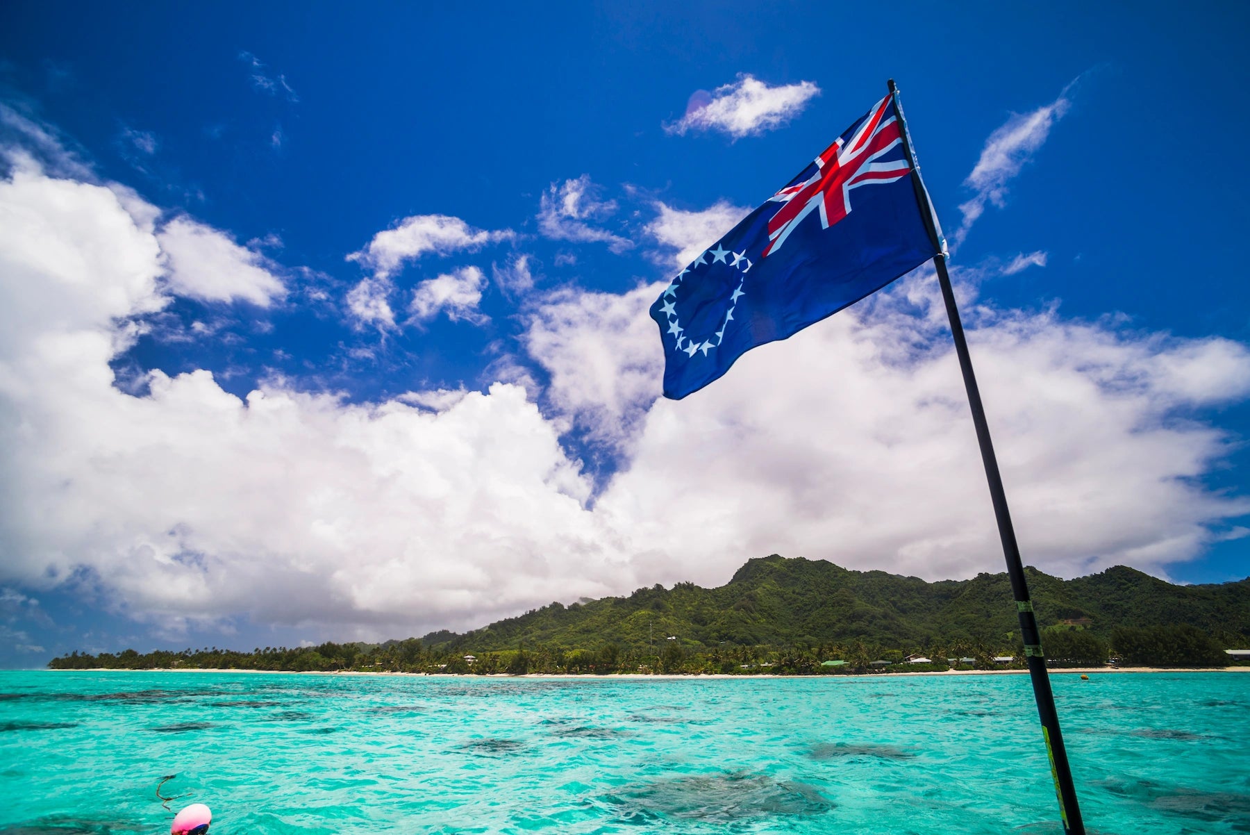 Die Flagge der Cook Islands vor dem Meer und einer grünen Insel.