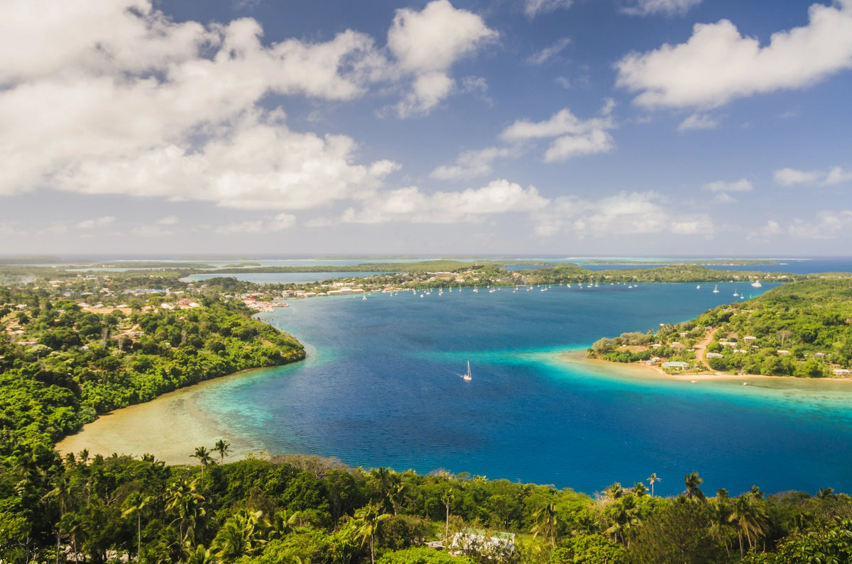 Luftaufnahme einer tiefblau schimmernden Meeresbucht auf Tonga, umgeben von dicht bewaldeten, grünen Inseln und Küstenstreifen unter leicht bewölktem Himmel.