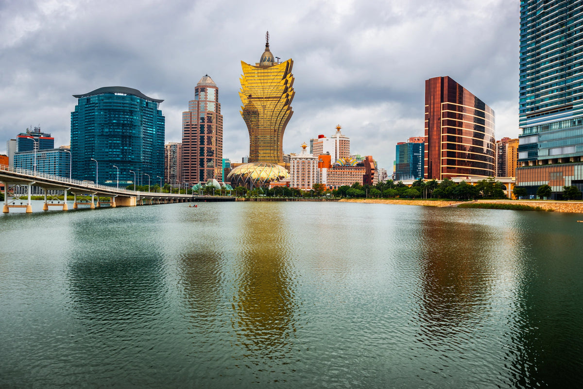 Blick über eine Wasserfläche auf die Skyline von Macau mit dem auffälligen goldene Hotelgebäude Grand Lisboa im Zentrum unter bedecktem Himmel.