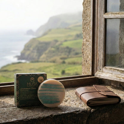 Der feste Conditioner Hanf von MUSA in seiner grünen Verpackung steht neben einem Leder-Notizbuch auf einem steinernen Fenstersims mit Blick auf eine neblige Küstenlandschaft.