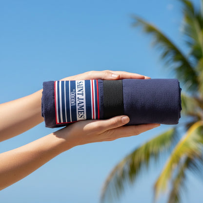 Zwei Hände halten das kompakt gerollte XXL-Strandtuch von OBABA in Dunkelblau mit dem gestreiften Saint James Logo vor einem strahlend blauen Himmel mit Palme.