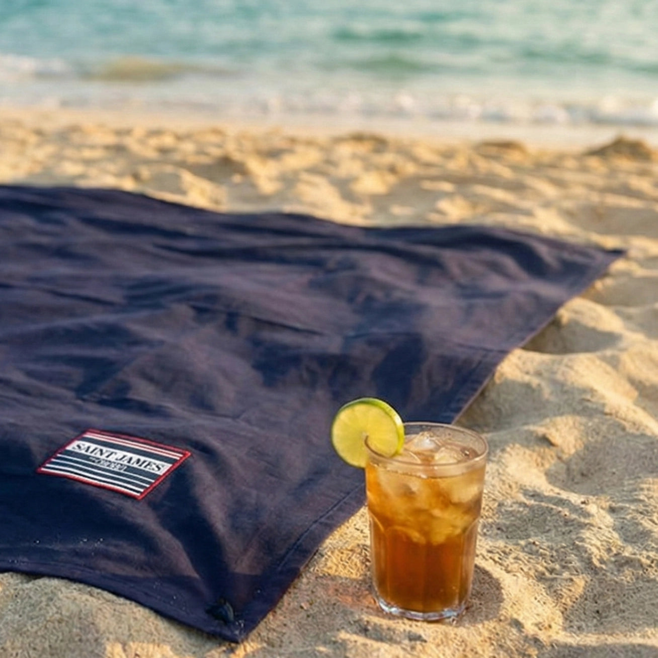 Ein ausgebreitetes, dunkelblaues XXL-Strandtuch von OBABA aus Bio-Baumwolle am Sandstrand, daneben steht ein Eistee mit Limette – perfekt für den nächsten Sommerurlaub.