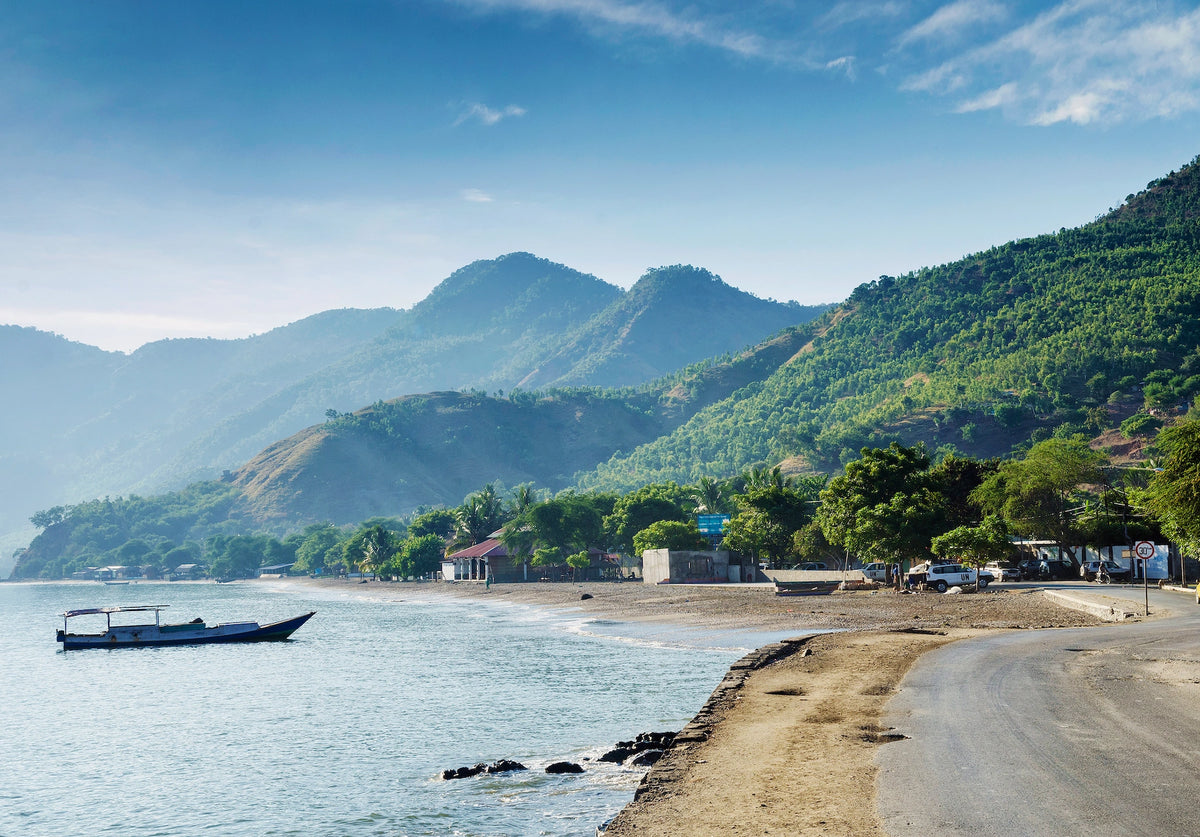 Küstenszene in Osttimor mit einer asphaltierten Straße direkt am Strand, einem kleinen Holzboot im ruhigen, blauen Wasser und dicht bewaldeten, grünen Bergen im Hintergrund.