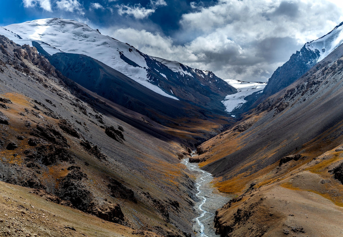 Imposantes, steiles Gebirgstal in Pakistan mit einem schmalen Schmelzwasserbach in der Sohle, flankiert von kargen, steinig-braunen Hängen und schneebedeckten Gipfeln unter leicht bewölktem Himmel.