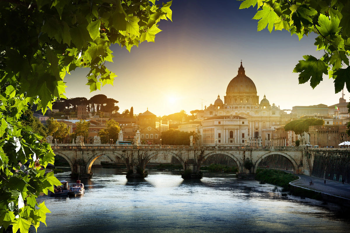 Blick über den Tiber und die Engelsbrücke auf den sonnenbeschienenen Petersdom im Vatikan, gerahmt von grünem Laub im Vordergrund.